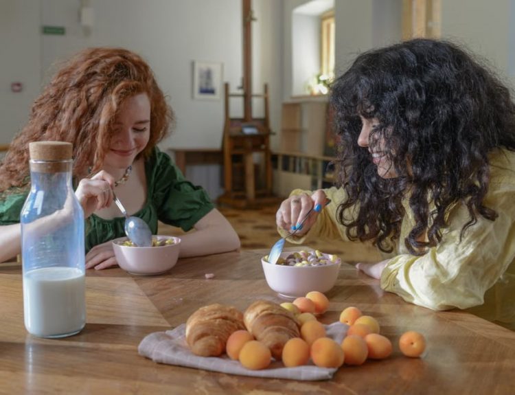 Two young women enjoying breakfast at home with cereal, croissants, and fruit around a wooden table.