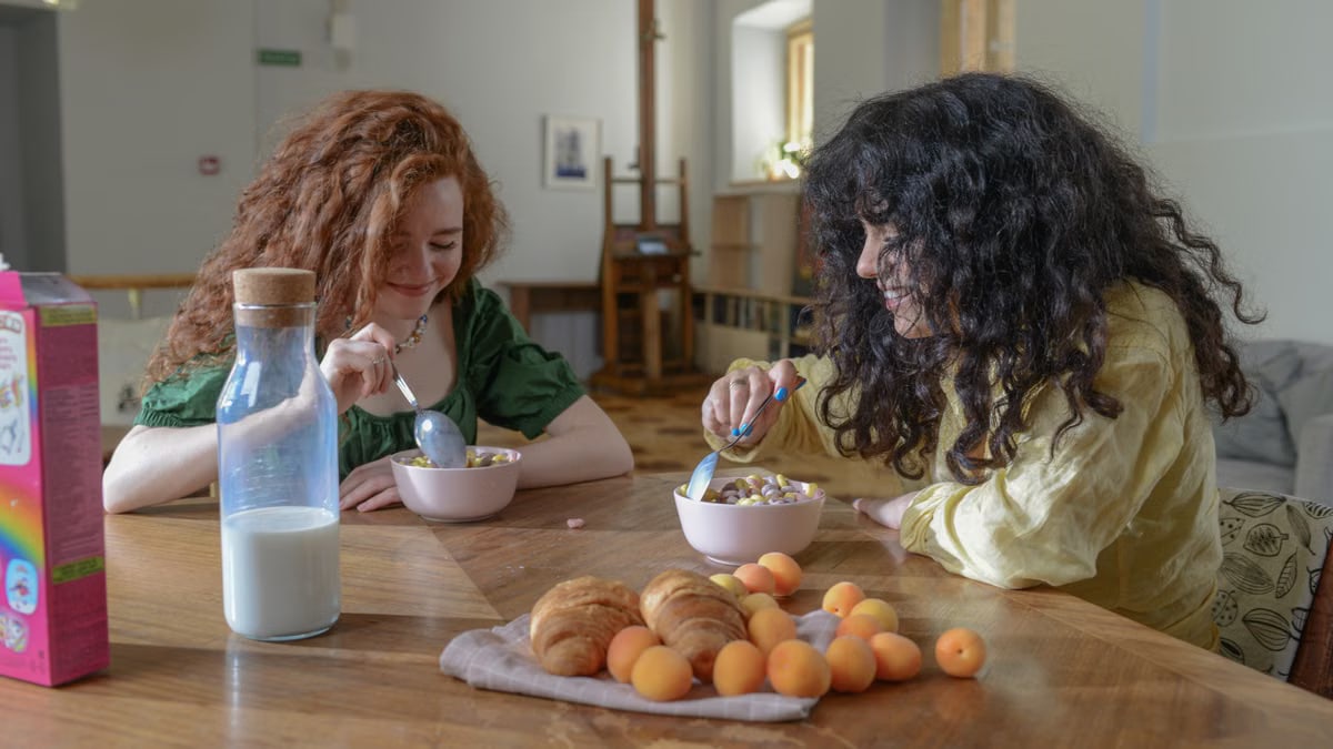 Two young women enjoying breakfast at home with cereal, croissants, and fruit around a wooden table.